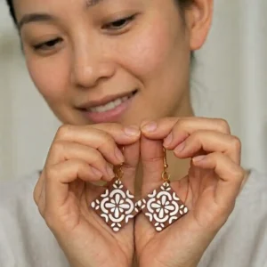 Deux mains tenant délicatement la paire de boucles d'oreilles losanges aux motifs blancs en relief.