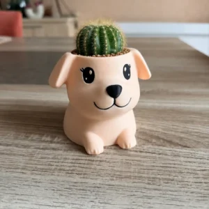 Pot en forme de chien mignon de la collection 'Verdanelle', couleur pêche mat, imprimé en 3D avec de jolies oreilles tombantes, accueillant une petite plante sur une table en bois.