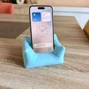 Support de téléphone bleu ciel en forme de mini canapé, imprimé en 3D, maintenant un iPhone 15 Pro Max sur une table.