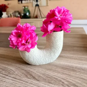 Vase double en forme de U blanc avec motifs de fleurs gravés en relief, imprimé en 3D, accueillant des roses d'un rose éclatant sur une table en bois.